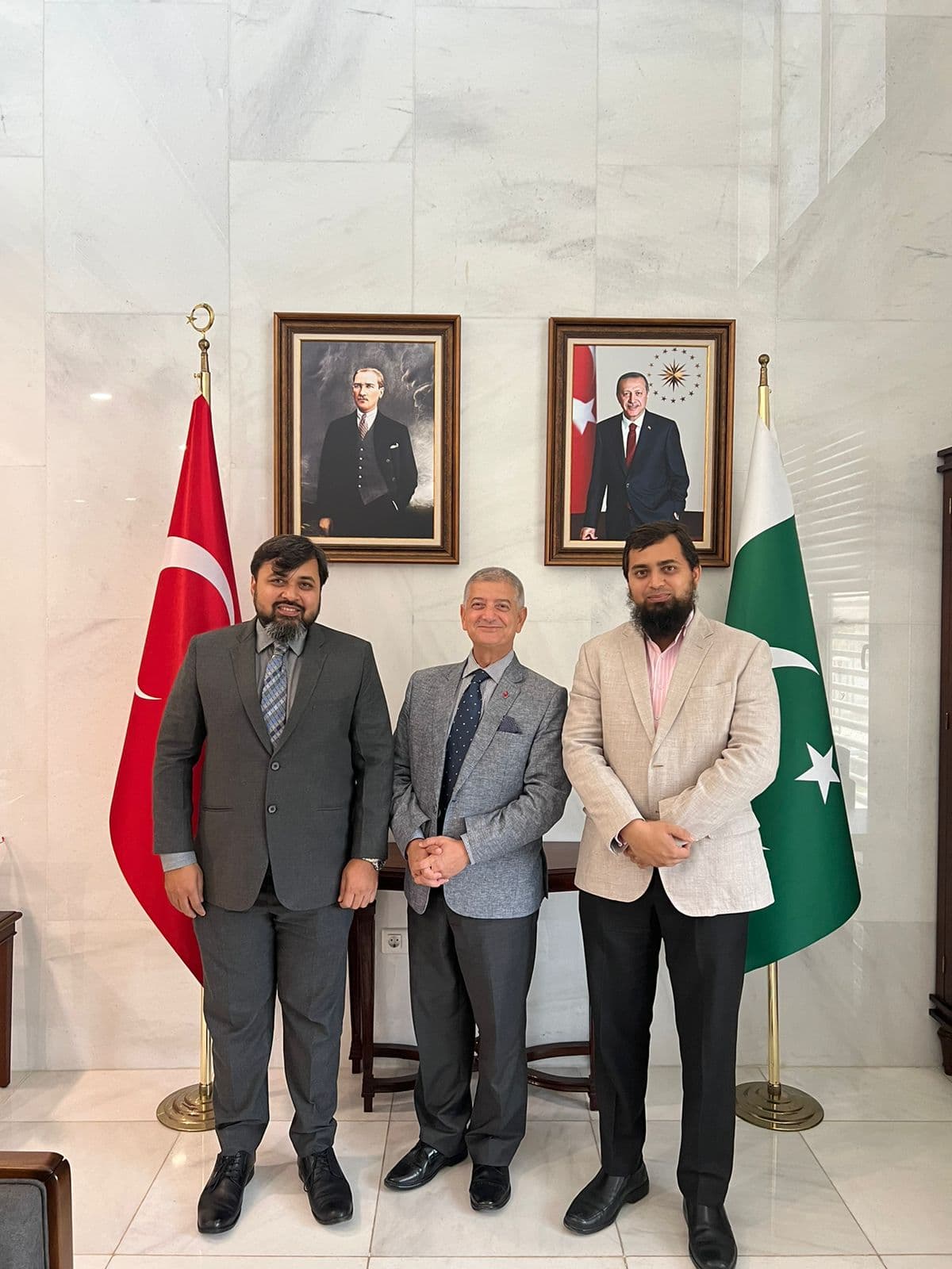 Engr. Atif Nazar Siddiqui, left, and Engr. Wasif Nazar Siddiqui, right, of ATIF NAZAR (Pvt.) Ltd. with Cemal Sangu, Consul General of Türkiye, centre, standing between Turkish and Pakistani flags